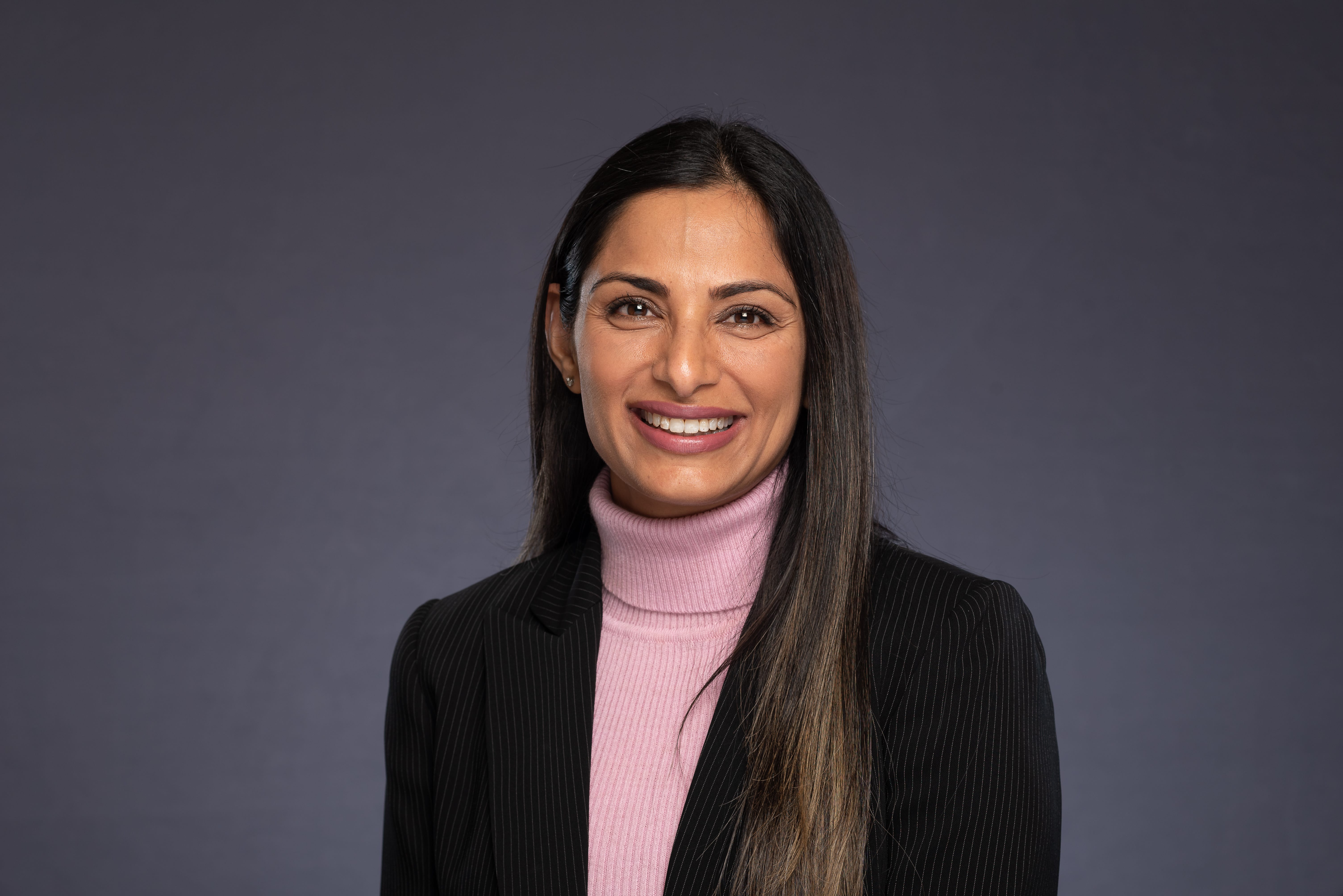 Professional headshot of a woman in a pink turtleneck and black blazer — dark studio background
